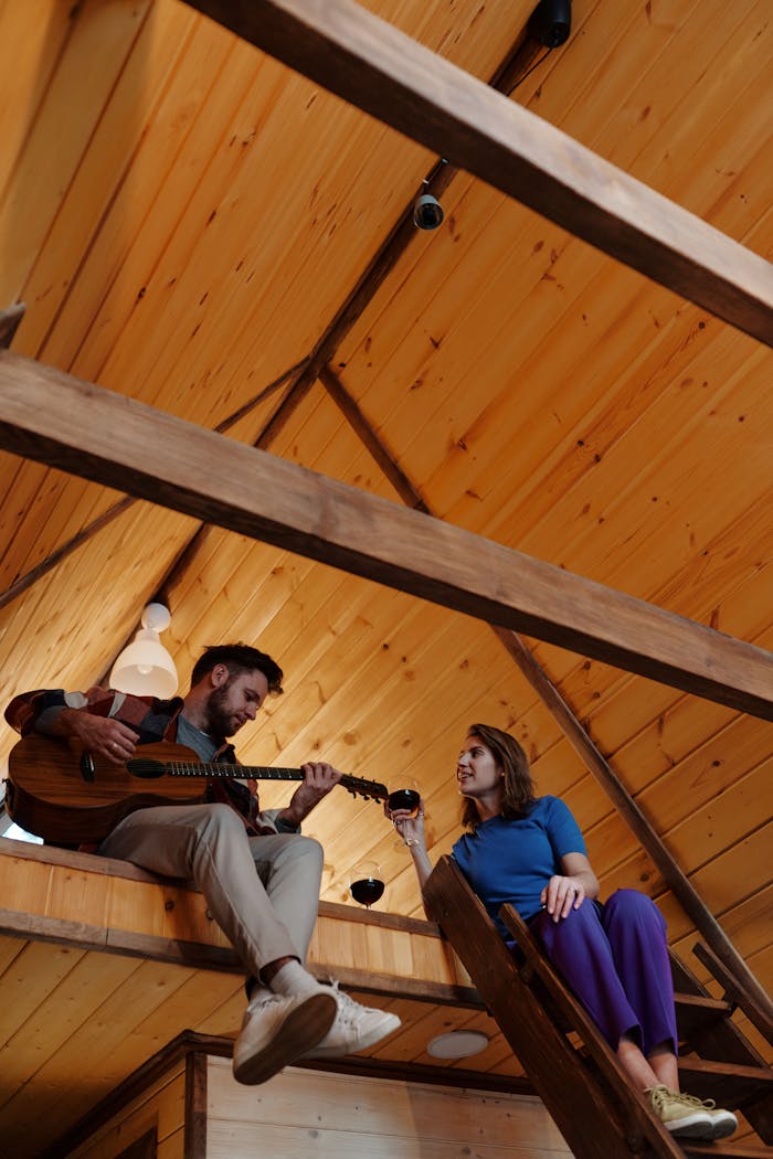 A couple enjoys playing guitar and sharing a warm drink in a wooden cabin loft.